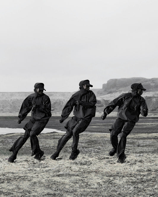 Three models in black techwear jackets and pants running outdoors in a rocky landscape