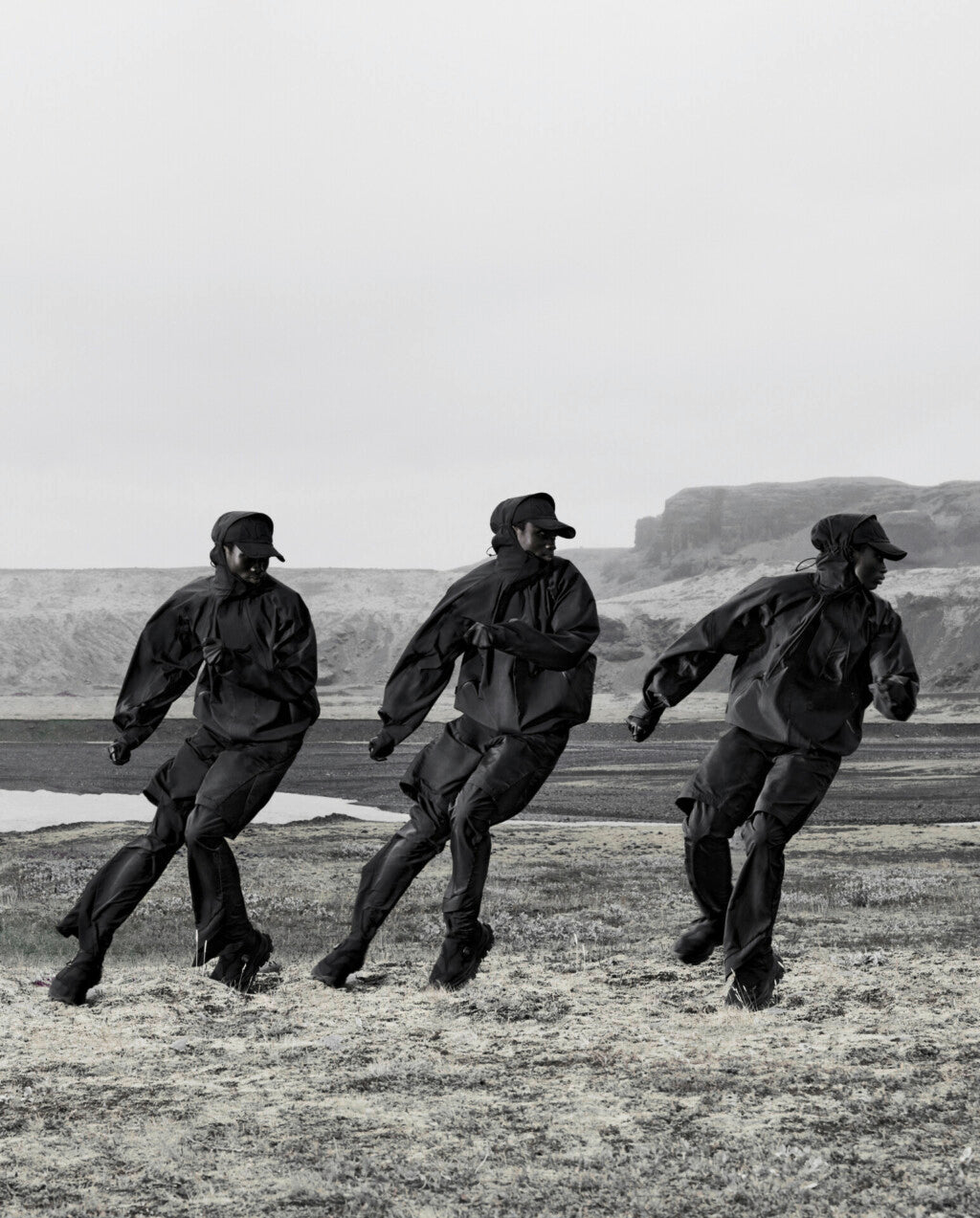 Three models in black techwear jackets and pants running outdoors in a rocky landscape